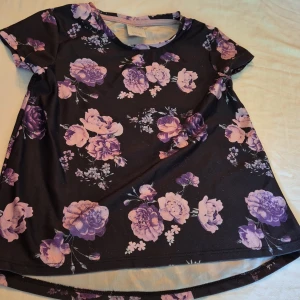 Svart blommig topp från KappAhl - Säljer en superfin svart topp från KappAhl med lila och rosa blommor. Toppen har korta ärmar och är perfekt för både vardag och fest. Materialet är mjukt och bekvämt, vilket gör den skön att ha på sig hela dagen. Passar perfekt till jeans eller kjol!