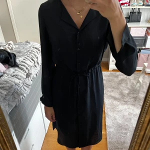 Svart långärmad klänning - Säljer en elegant svart långärmad klänning. Den har en knäppning framtill och ett knytband i midjan för en fin passform. Den är köpt från vila och är i storlek 36💫