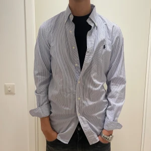Randig skjorta från Ralph Lauren - Säljer en snygg och stilren randig skjorta från Ralph Lauren i storlek S. Skjortan är vit med tunna blå ränder och har en broderad logga på bröstet. Den är långärmad och har knappar hela vägen ner. Perfekt för både vardag och fest!