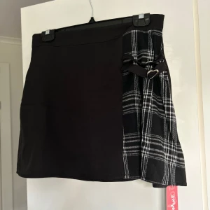 Svart kortkjol med rutigt mönster - Säljer en superfin svart kortkjol med en cool rutig detalj på sidan. Kjolen har en litet spänne som ger den en extra touch. Perfekt för både vardag och fest! 🖤