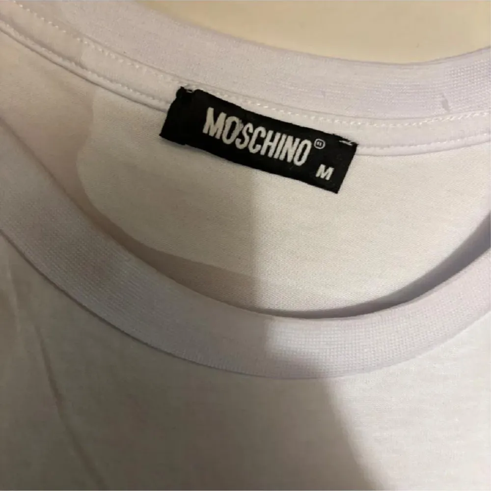 Säljer en supercool vit t-shirt från Moschino med ett stort tryck av en nallebjörn som bär en Moschino-keps. T-shirten har korta ärmar och är i bomull, perfekt för en avslappnad och trendig look. Passar perfekt till både jeans och shorts!. T-shirts.