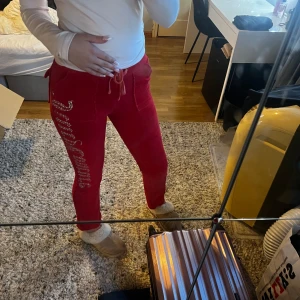 Röda mjukisbyxor med paljetter - Säljer ett par snygga röda Juicy couture där det står Juicy Couture längs sidan med paljetter. De har en bekväm passform och snörning i midjan. Perfekta för en avslappnad dag hemma eller när du vill glittra lite extra!