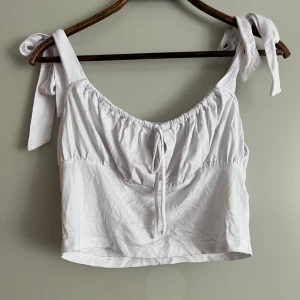 Vit croppad topp med knytband - Säljer en superfin vit croppad topp med knytband på axlarna. Toppen har en söt knytdetalj framtill och är perfekt för sommardagar. Den är ärmlös och har en luftig passform som gör den bekväm att bära. Perfekt för en avslappnad dag på stan eller en sommarfest!