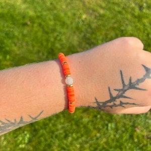 Orange armband med pärlor - Säljer ett snyggt armband i en härlig orange färg. Armbandet har små, runda pärlor och en större, glittrig pärla i mitten som ger det en extra touch. Perfekt för sommarens fester eller bara för att liva upp vardagen!