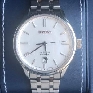 Säljer en elegant Seiko Presage Automatic klocka i nyskick. Den har en stilren vit urtavla med silverdetaljer och datumvisning. Boetten är i rostfritt stål och baksidan visar den automatiska mekanismen med 23 juveler. Armbandet är också i rostfritt stål och ger en klassisk look. Perfekt för både vardag och fest!