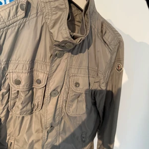 Beige jacka från Moncler - Säljer en snygg beige jacka från Moncler med långa ärmar och flera praktiska fickor framtill. Jackan har knappar och dragkedja för stängning samt en stilren krage. Perfekt för vår och höst! Skick 9/10 knappt använd