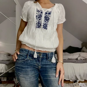 Vit blus med blå broderi - Charmig vit blus med blått broderi framtill. Blusen har korta ärmar och en knytning vid halsen med tofsar. Perfekt för en avslappnad och stilren look.