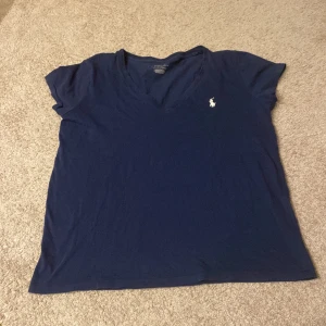 Mörkblå t-shirt från Ralph Lauren - Säljer en mörkblå t-shirt från Ralph Lauren med v-ringning och korta ärmar. Den har en liten vit logga broderad på bröstet. Perfekt för en avslappnad stil. Passar xs