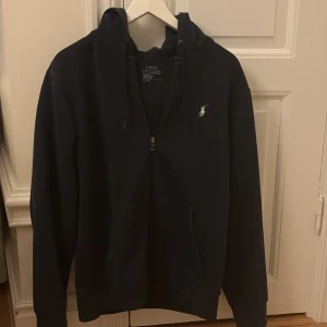 Mörkblå hoodie från Polo Ralph Lauren - Säljer en stilren mörkblå hoodie från Polo Ralph Lauren med dragkedja och det klassiska logotypbroderiet på bröstet. Tröjan har långa ärmar och en praktisk huva med snörning. Perfekt för en avslappnad look.