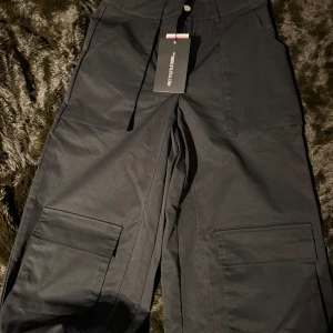 Svarta cargobyxor från PrettyLittleThing - Snygga svarta cargobyxor från PrettyLittleThing med praktiska fickor på sidorna. Byxorna har en loose passform och är perfekta för en avslappnad stil. De är högmidjade och har en stilren design som passar till många olika outfits.