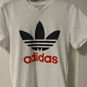 Vit t-shirt från Adidas - Snygg vit t-shirt från Adidas med det klassiska trefoil-loggan i svart och röd text. Perfekt för en sportig look eller avslappnad stil. Kortärmad och tillverkad i mjukt material för extra komfort.