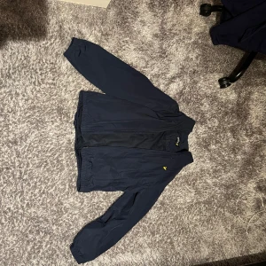 Mörkblå jacka från Lyle & Scott - Säljer en stilren mörkblå jacka från Lyle & Scott. Jackan har långa ärmar och en dragkedja framtill. Perfekt för en casual look. Passar 14-15 åringar enligt storleksguide. Nypris: 699kr