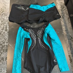 Svart och turkos gymnastikdräkt med shorts - Snygg gymnastikdräkt i svart och turkos med matchande shorts. Dräkten har långa ärmar och är dekorerad med glittriga detaljer. Perfekt för träning eller tävling. Det passar barn mellan åldern 10-11 år 