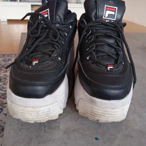 Svarta sneakers från Fila - Snygga svarta sneakers från Fila med tjock vit sula och snörning. Skorna har en sportig look med Fila-loggan i rött och vitt på sidan och tungan. Perfekta för en casual stil. Använda max 15.man ser dom är använda men ej trasiga på något sätt storlek 38 1/2