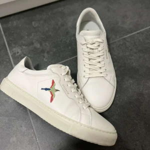 Vita sneakers från Axel Arigato - Snygga vita sneakers från Axel Arigato med broderad fågel på sidan. Skorna har en klassisk design med snörning och är tillverkade i skinn. Perfekta för en stilren look.