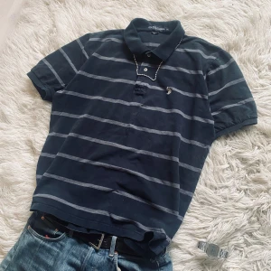 Mörkblå randig pikétröja från U.S. Polo Assn. - Säljer en snygg mörkblå pikétröja från U.S. Polo Assn. med ljusgrå ränder. Tröjan har en klassisk krage och korta ärmar, perfekt för en avslappnad stil. Den är tillverkad i bomull för en bekväm passform. Nyskick med inga defekter!