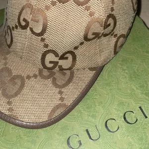 Beige keps från Gucci - Snygg beige keps från Gucci med det ikoniska GG-mönstret i brunt. Kepsen har en klassisk design med böjd skärm och justerbar passform. Perfekt för att ge din outfit en lyxig touch.
