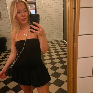 Svart axelbandslös klänning - Snygg svart axelbandslös klänning med volangdetalj i nederkant. Perfekt för en kväll ute eller en speciell tillställning. Enkel och stilren design som passar till många olika accessoarer.