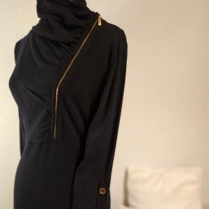 Svart klänning med dragkedja från Flash Woman - Snygg svart klänning från Flash Woman med unik diagonal dragkedja och hög krage.  Tillverkad av en mjuk blandning av viskos och nylon.  Perfekt för en stilren look