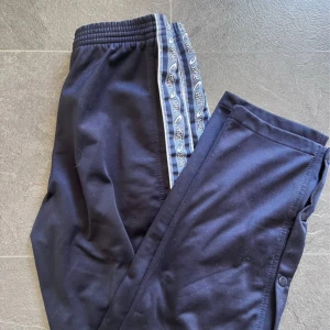 Blåa träningsbyxor från Asics - Snygga blåa vintage träningsbyxor från Asics med band i midjan och knappar längs sidorna. Byxorna har en dekorativ rand med mönster längs benen och vita detaljer vid bensluten. 