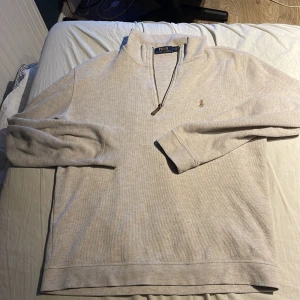 Beige tröja från Ralph Lauren - Säljer en stilren beige tröja från Ralph Lauren med dragkedja och broderad logga på bröstet. Perfekt för en avslappnad och snygg look. Tröjan har långa ärmar och en bekväm passform.