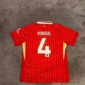 Liverpool FC | 24/25 | Size: L | Hemmatröja | Virgil | Ny, oanvänd - Liverpools hemmatröja för nuvarande säsong 2024-2025. Virgil, nr 4 på ryggen. Stadium/fan edition. Ny & oanvänd. Storlek large. Tröjan är en nyproducerad reprint i utmärkt kvalitet. Perfekt för alla Liverpool fans där ute!⚽  Skickas 1-3 dagar efter mottagen betalning. Har du frågor/funderingar är det bara att höra av dig! :)