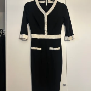 Svart klänning med vita detaljer - Elegant svart klänning med vita kontrastdetaljer och knappar framtill. Klänningen har trekvartsärmar och en v-ringad design. Perfekt för en stilren look.