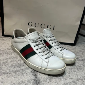 Vita sneakers från Gucci - Snygga vita sneakers från Gucci med klassiska gröna och röda ränder på sidan. Skorna har snörning och en grön detalj på hälen. Perfekta för en stilren look.