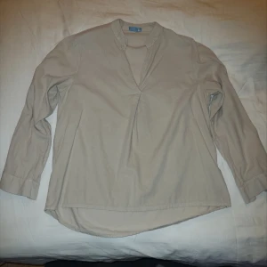 Beige blus med lång ärm - Snygg beige blus med lång ärm och en stilren design. Perfekt för en elegant look. Blusen har en diskret v-ringning och är tillverkad i ett manchestertyg. 