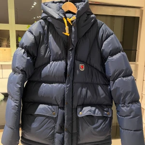 Blå dunjacka från Fjällräven - Säljer en blå dunjacka från Fjällräven Expedition Series. Jackan har en pufferdesign med långa ärmar och en praktisk huva. Den är utrustad med dragkedja och knappar framtill samt två fickor med lock. Perfekt för kalla dagar! 