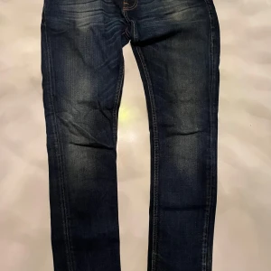Nudie jeans - Snygga mörkblå jeansbyxor med en klassisk design. De har en smal passform och är perfekta för en stilren look. Byxorna har en traditionell femficksmodell och en knappgylf.
