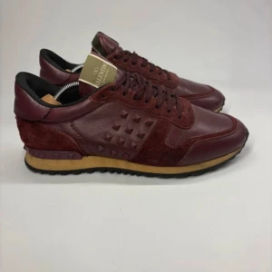 Vinröda sneakers från Valentino - Snygga vinröda sneakers från Valentino med en kombination av skinn och mocka. Skorna har en unik design med pyramidformade nitar på sidorna och en kontrasterande beige sula. Perfekta för att ge din outfit en lyxig touch. Skick 9/10 en nit saknas  