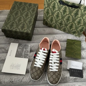 Gucci sneakers med monogrammönster - Snygga Gucci sneakers med det ikoniska monogrammönstret i beige och brunt. Skorna har vita snören och en röd och blå randdetalj på sidan. Insidan är rosa och sulan är vit med ett präglat mönster. Perfekta för en stilren look. Skorna är endast testade och säljs eftersom att de inte passade! Pris kan diskuteras vid snabb affär!