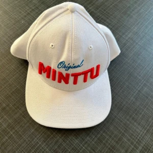 Vit keps från MINTTU - Snygg vit keps med broderad text 'Original MINTTU' i rött och blått framtill. Justerbar passform med plastspänne baktill. Perfekt för en avslappnad stil.