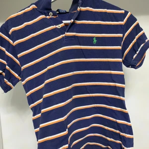 Randig pikétröja från Ralph Lauren - Säljer en snygg randig pikétröja från Ralph Lauren i blått, vitt och orange. Tröjan har korta ärmar och en klassisk krage med knappar. Perfekt för en avslappnad stil.