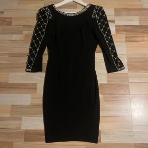 Svart klänning med pärldetaljer - Elegant svart klänning med trekvartsärm och pärldetaljer på ärmarna. Klänningen har en djup v-ringning i ryggen och en dragkedja för en stilren look. Perfekt för en kväll ute eller en speciell tillställning.