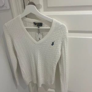 Vit stickad tröja från Polo Ralph Lauren - Säljer en stilren vit stickad tröja från Polo Ralph Lauren med v-ringning och det klassiska logotypbroderiet på bröstet. Tröjan har långa ärmar och en ribbad struktur som ger en elegant look. Perfekt för kyligare dagar.