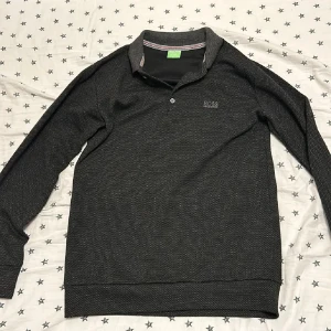 Svart pikétröja från Hugo Boss - Snygg svart pikétröja från Hugo Boss med diskret mönster och klassisk krage. Tröjan har knappar framtill och är perfekt för en stilren look. Passar bra till både jeans och chinos.