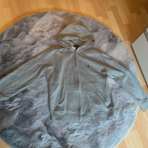 Grå hoodie från Ralph Lauren - Säljer en stilren grå hoodie från Ralph Lauren med dragkedja och broderad logga på bröstet. Perfekt för en avslappnad look. Den har en bekväm passform och praktiska fickor framtill. Jätte bra skick. Köpt på Kids brandstore så är i barnstorlek XL men passar 12-16 år. Den är 100% äkta.