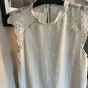 Vit ärmlös blus med spetsdetaljer - Säljer en elegant vit blus med spetsdetaljer vid axlarna och ett diskret prickigt mönster. Blusen är ärmlös och har en knapp i nacken för enkel påklädning. Perfekt för en stilren look.