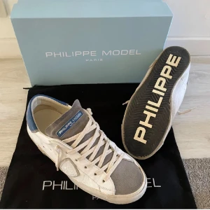 Philipe model skor (helt nya) - Snygga sneakers från Philippe Model i vitt och grått med blå detaljer. Skorna har snörning och en stilren design med märkets logga på sulan. Perfekta för en avslappnad och trendig look. Dem är bara använda ett fåtal gånger