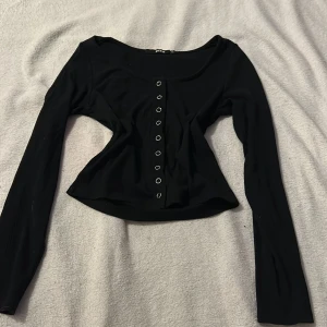 Svart ribbad topp från Bik Bok - Säljer en svart ribbad topp från Bik Bok i storlek XS. Toppen har långa ärmar och knappar framtill, vilket ger en stilren look. Perfekt för att matcha med dina favoritjeans eller kjol.