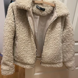 Beige teddyjacka från Zara - Mysig beige teddyjacka från Zara med dragkedja framtill. Perfekt för kyliga dagar med sin fluffiga och varma design. Jackan har långa ärmar och en bekväm passform.