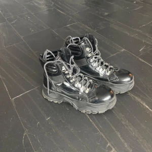 Buffalo kängor, lack i stl 40 svarta med snörning - Snygga svarta Buffalo kängor, stl 40, med robust snörning och kraftig sula. Perfekta för en tuff look och passar bra till höst- och vinterväder. Kängorna har en glansig finish (lack) och metalliska detaljer vid snörningen. De är i fint skick.