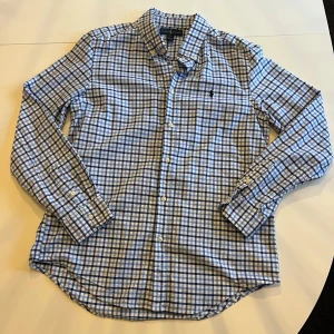 Ralph Lauren skjorta - Snygg blårutig skjorta från Ralph Lauren med klassisk krage och broderad logga på bröstet. Skjortan har långa ärmar och knappar framtill. Perfekt för en stilren look.