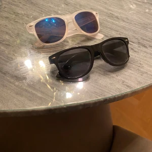 Två par solglasögon - Säljer två par solglasögon. Ett par är svart med mörka linser och det andra är vitt med blåtonade linser. Perfekta för soliga dagar och ger en stilren look. Båda har en klassisk form som passar de flesta ansikten.