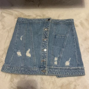 Blå jeanskjol med knappar - Snygg blå jeanskjol från only som även går att ha som topp med slitna detaljer och knappar framtill. Perfekt för en avslappnad stil. Kjolen har en kort längd och en klassisk jeanslook. 