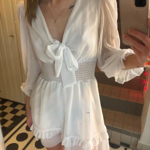 Vit klänning med knyt och volang - Charmig vit klänning med knyt framtill och volangdetaljer. Den har långa ärmar och en smockad midja som ger en fin passform. Perfekt till studenten