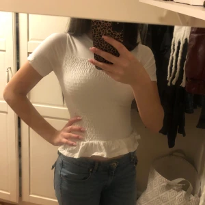 Vit topp från Gina Tricot - Säljer en söt vit topp från Gina Tricot med korta ärmar och volangdetalj vid nederkanten som aldrig är använd. Toppen har en smockad design som ger en fin passform. Perfekt för en stilren och bekväm look💗💗