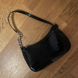 Svart axelväska med krokodilmönster - Snygg svart axelväska i syntet med krokodilmönster och en stilren design. Väskan har en dragkedja och en kedjedetalj på axelremmen som ger en extra touch. Perfekt för att lyfta din outfit!
